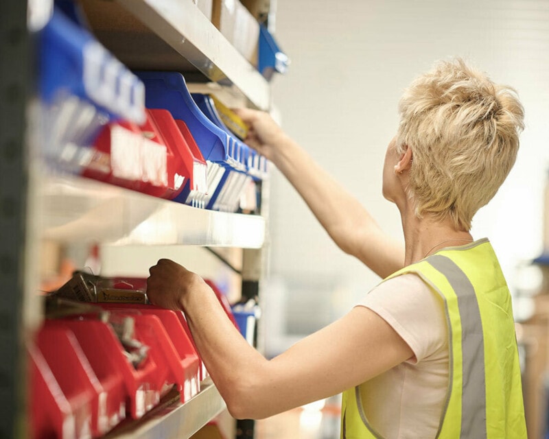 A warehouse worker with short blond hair reaches upwards to an item on a shelf. She is wearing a hi vis vest and is facing away from the camera.