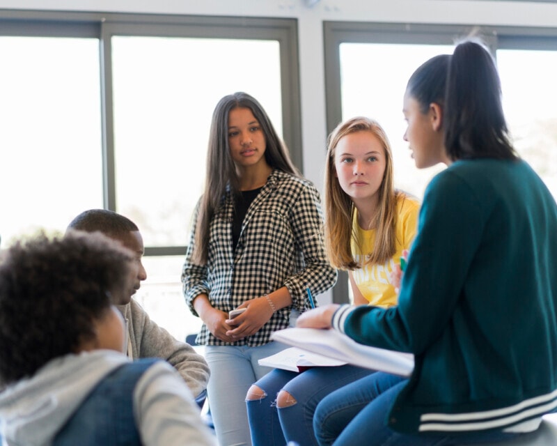 Teenage students sitting on and around a table in a classroom, talking. They are all facing one girl with a pony tail, who is speaking.