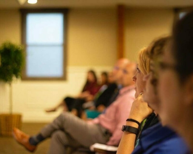 Side view of a line of sitting people listening to a presentation in a room with dimmed lights.