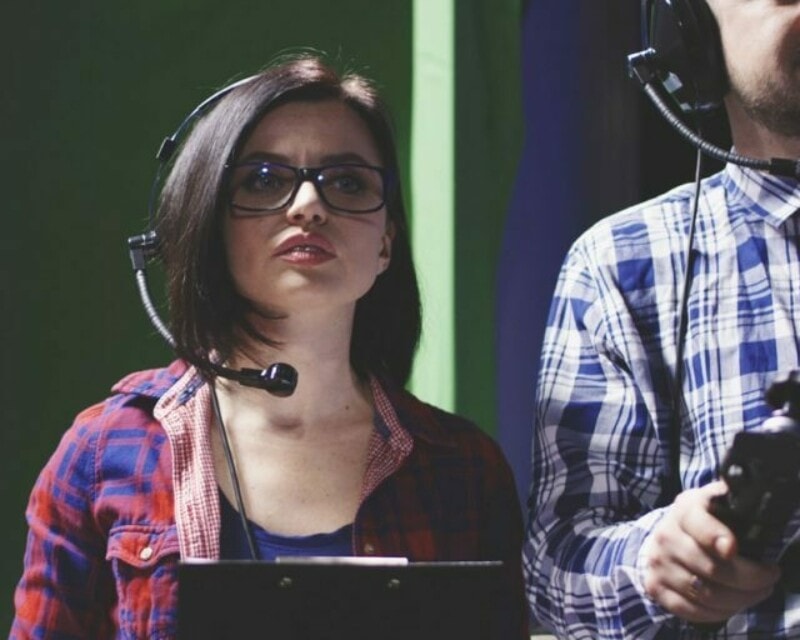 Woman at work on a TV set is wearing a headset, looking ahead just over the camera. A man operating filming equipment stands at her left, his image is cropped.