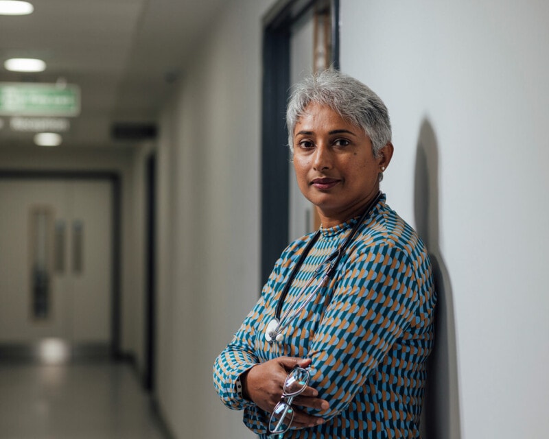 A middle aged woman with brown skin and short grey hair stands in a hospital corridor with her arms crossed at her chest. She has a stethoscope around her neck.