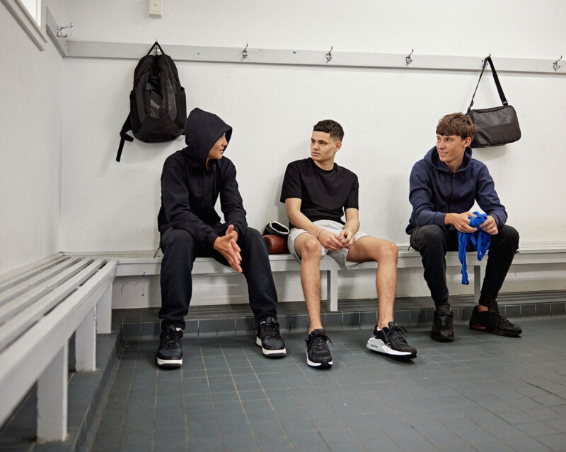 Three young people seated on a bench in a changing room, talking with one another.