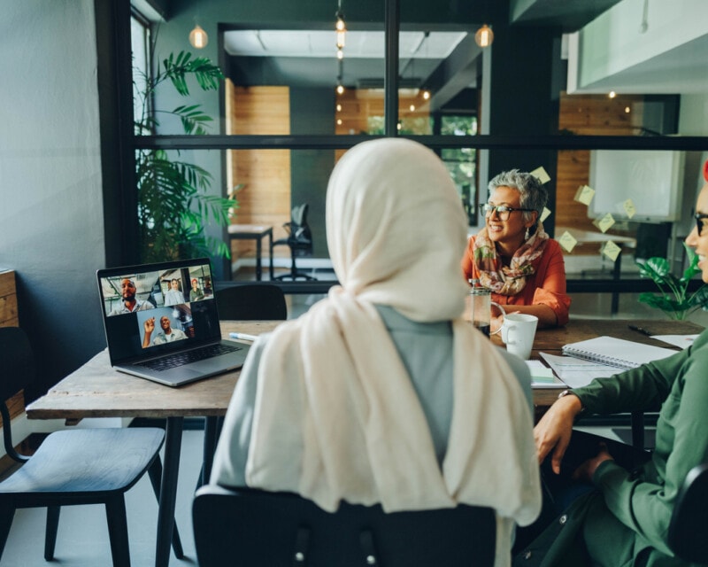 Three women in head covering at a board room in an online meeting with coworkers on the screen.