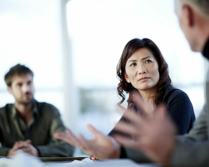 An Asian Australian woman is sitting in a meeting listening to a male colleage, with another male colleage next to her. She is listening intently.