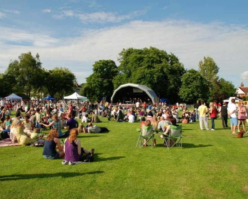 Outdoor community event with a large group of people on a lawn sitting around in front of a stage. There are trees in the background and blue sky.